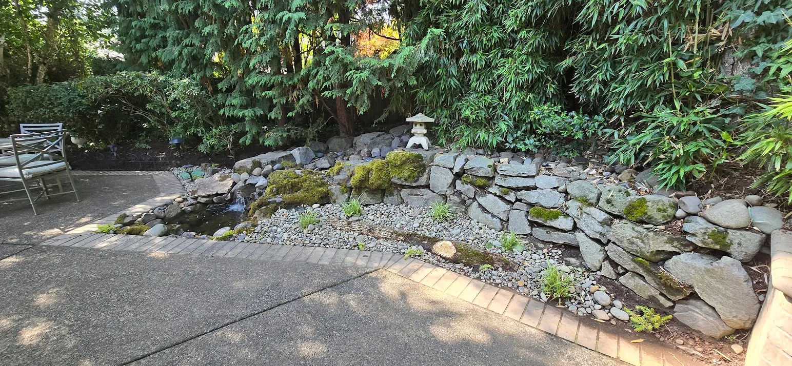 Stacked-stone waterfall spilling into a pebble pond with fresh river rock and new grasses in the same corner